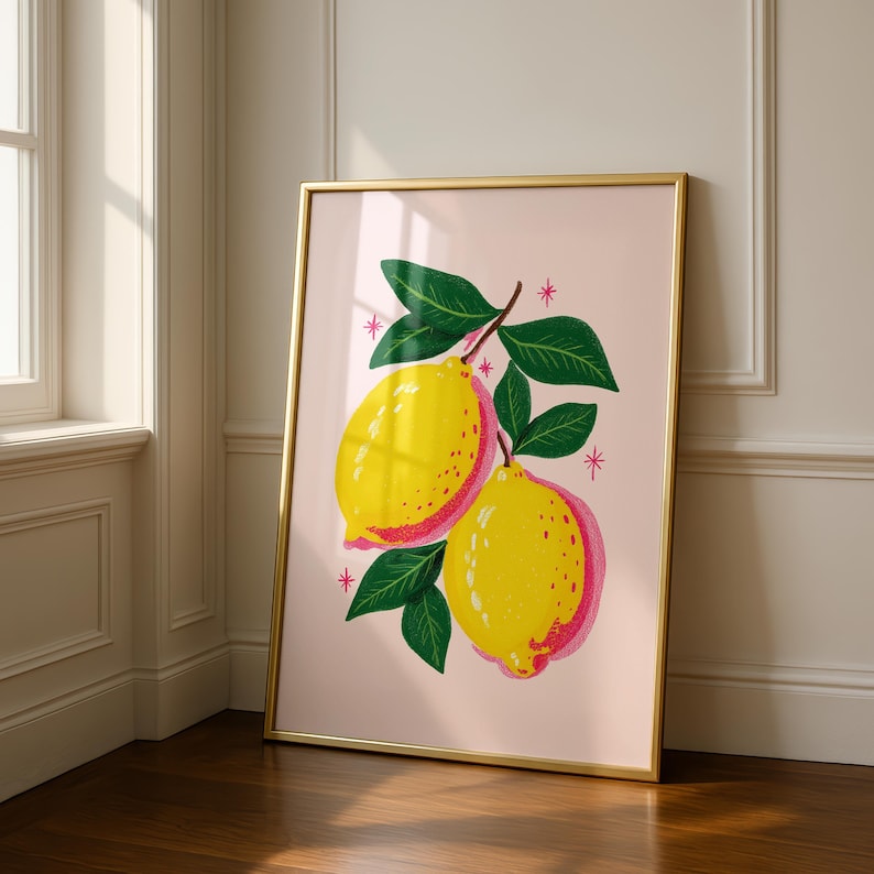 A gold framed print of two yellow and pink lemons on a branch with green leaves, on a light pink background with small pink starbursts, leaning against a white wall and wood floor.