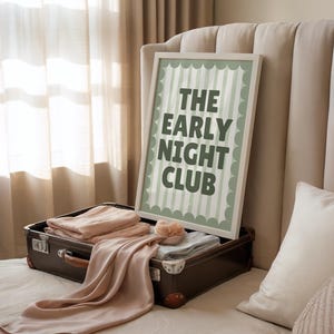May include: A framed print with the text "THE EARLY NIGHT CLUB" in a white frame, resting on an open brown suitcase filled with folded peach-colored textiles. A peach-colored scarf drapes over the suitcase. The background includes a beige headboard and curtains.