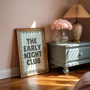 May include: A framed print with the text "THE EARLY NIGHT CLUB" in dark green letters. The frame is gold with a scalloped edge. The print is leaning against a wall next to a small, light blue cabinet with a lamp and a vase of pink flowers.