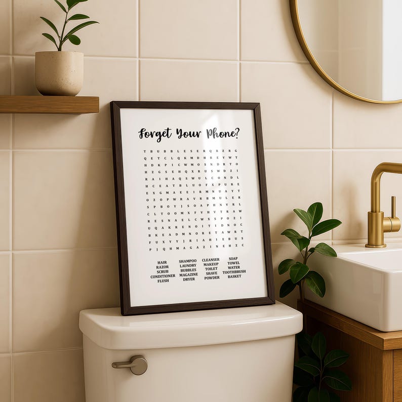 A dark wood-framed print with "Forget Your Phone?" and a word search puzzle, sitting on the tank of a white toilet, with a plant on a shelf and a round mirror.