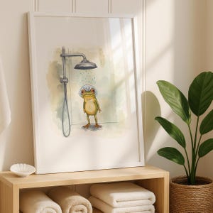 May include: A framed art print featuring a whimsical illustration of a frog taking a shower. The frog, in shades of green and yellow, stands under a silver shower head. The background has soft watercolor washes of green and blue. The print is in a white frame.
