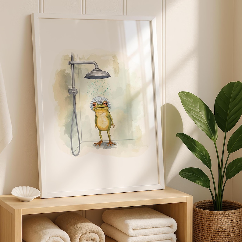 May include: A framed art print featuring a whimsical illustration of a frog taking a shower. The frog, in shades of green and yellow, stands under a silver shower head. The background has soft watercolor washes of green and blue. The print is in a white frame.