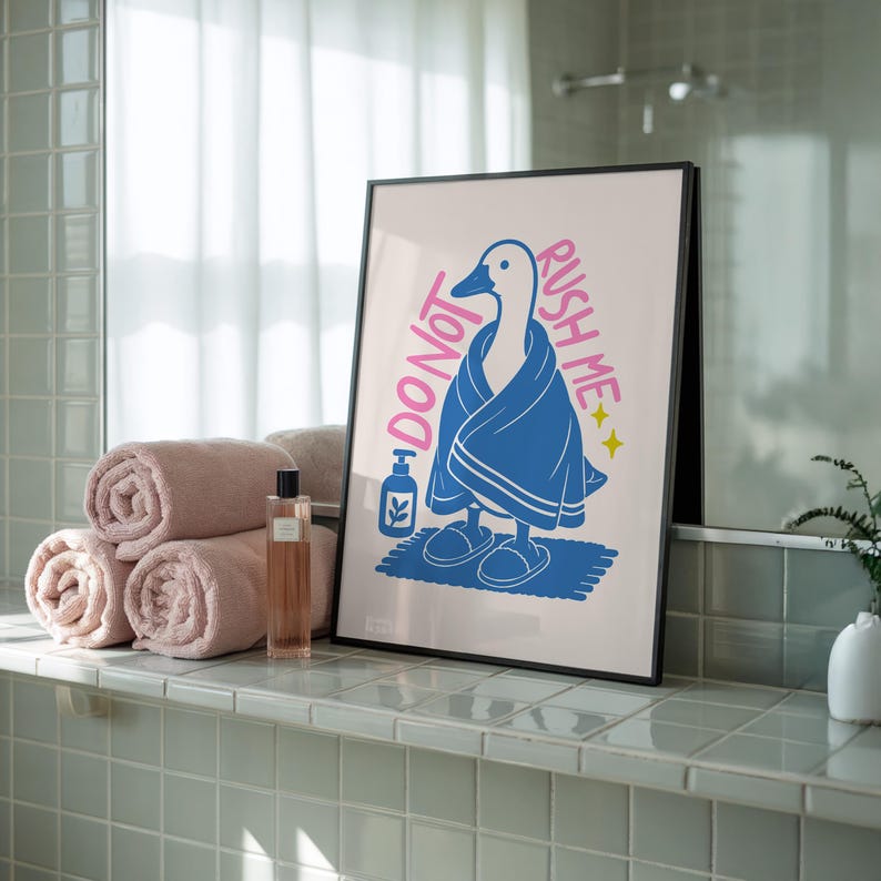 A framed print of a goose in a blue towel and slippers, with "DO NOT RUSH ME" text, rests on a tiled shelf next to rolled pink towels and a perfume bottle.