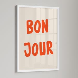 May include: A framed art print with the words "BON JOUR" in bold orange letters on a cream background. The print is encased in a white frame, creating a clean and modern aesthetic. The artwork is suitable for home decor.
