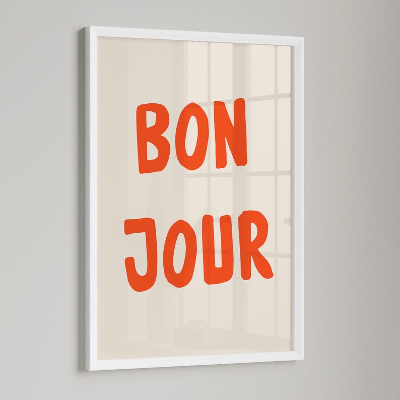 May include: A framed art print with the words "BON JOUR" in bold orange letters on a cream background. The print is encased in a white frame, creating a clean and modern aesthetic. The artwork is suitable for home decor.