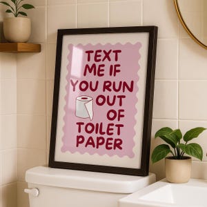 May include: A framed wall art print featuring a light pink scalloped background with dark red text that reads "TEXT ME IF YOU RUN OUT OF TOILET PAPER" and a small illustration of a white toilet paper roll centered between the lines of text.