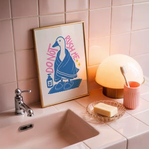A framed print of a goose in a blue towel and slippers, with "DO NOT RUSH ME" text, stands on a pink tiled counter next to a sink, soap, and toothbrush.