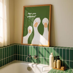 A framed print with a green background and three white geese, with the text "Are you pooping?" above them, sits on a bathroom counter next to a bathtub.