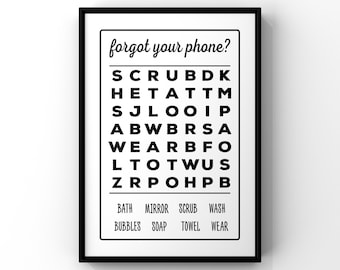 May include: A framed wall art print featuring a word search puzzle with the heading "forgot your phone?" in black text on a white background. Below the grid, the words "BATH", "MIRROR", "SCRUB", "WASH", "BUBBLES", "SOAP", "TOWEL", and "WEAR" are listed.