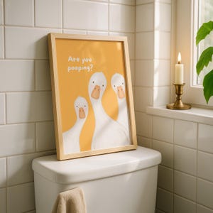 A framed print with a yellow background and three white geese, with the text "Are you pooping?" above them, leans against a toilet tank.