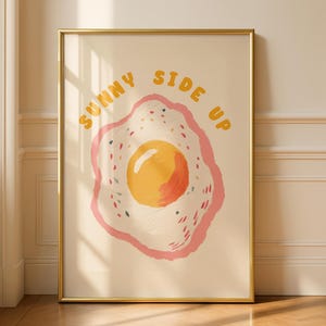 A framed print of a sunny-side-up egg with a pink and yellow border and the words "SUNNY SIDE UP" in yellow letters above it, leaning against a light beige wall.