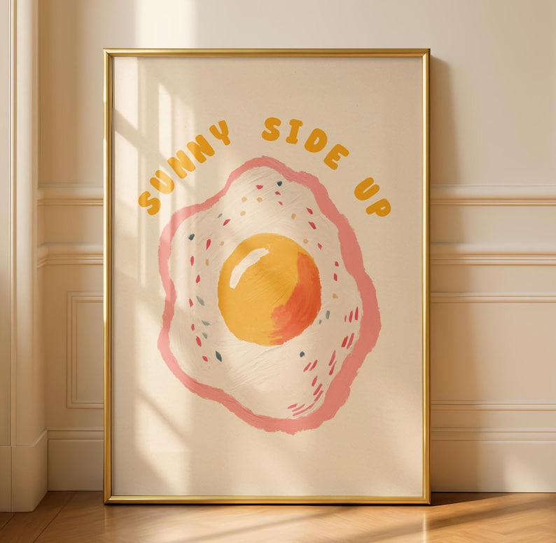 A framed print of a sunny-side-up egg with a pink and yellow border and the words "SUNNY SIDE UP" in yellow letters above it, leaning against a light beige wall.
