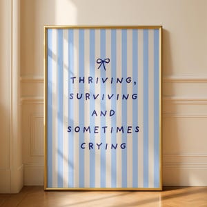 Puede incluir: Una impresión enmarcada con un borde dorado presenta un fondo a rayas azules y blancas. El texto en azul marino dice "Thriving, Surviving, and Sometimes Crying" con un lazo gráfico. La impresión se muestra contra una pared neutra.