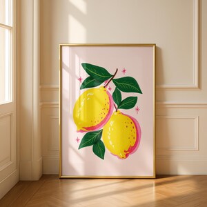 A gold framed print of two yellow and pink lemons on a branch with green leaves, on a light pink background with small pink starbursts, leaning against a white wall and wood floor.