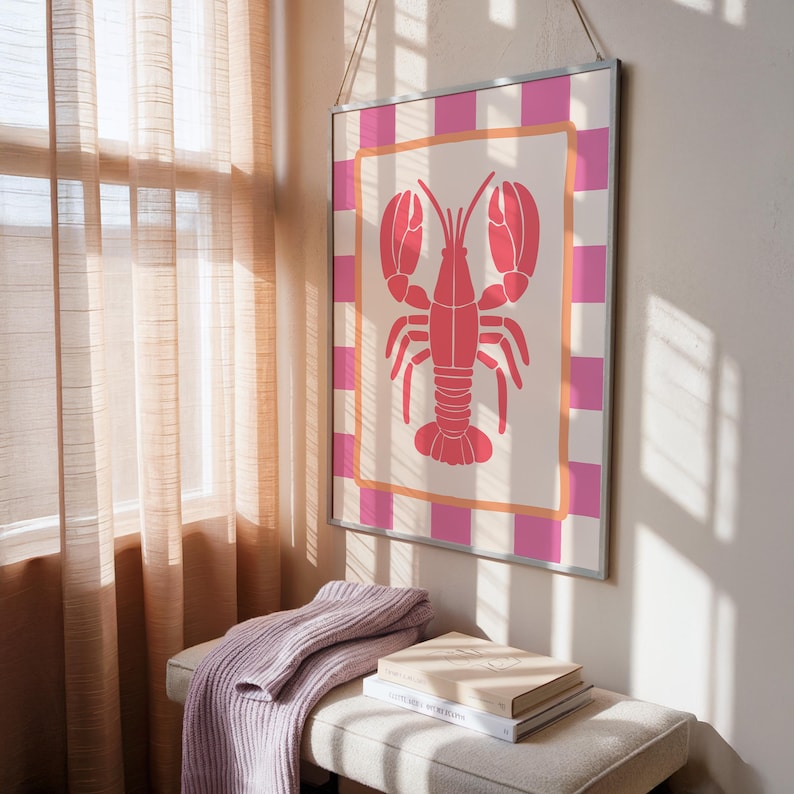 A framed print of a pink lobster on a cream background with a pink and white checkered border, hanging on a light-colored wall above a bench with a purple sweater and books.
