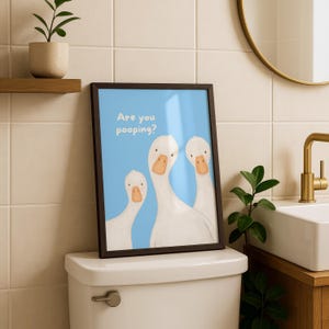 A framed print with a blue background and three white geese, with the text "Are you pooping?" above them, sits on a toilet tank.