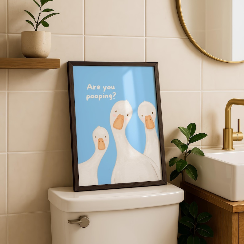 A framed print with a blue background and three white geese, with the text "Are you pooping?" above them, sits on a toilet tank.