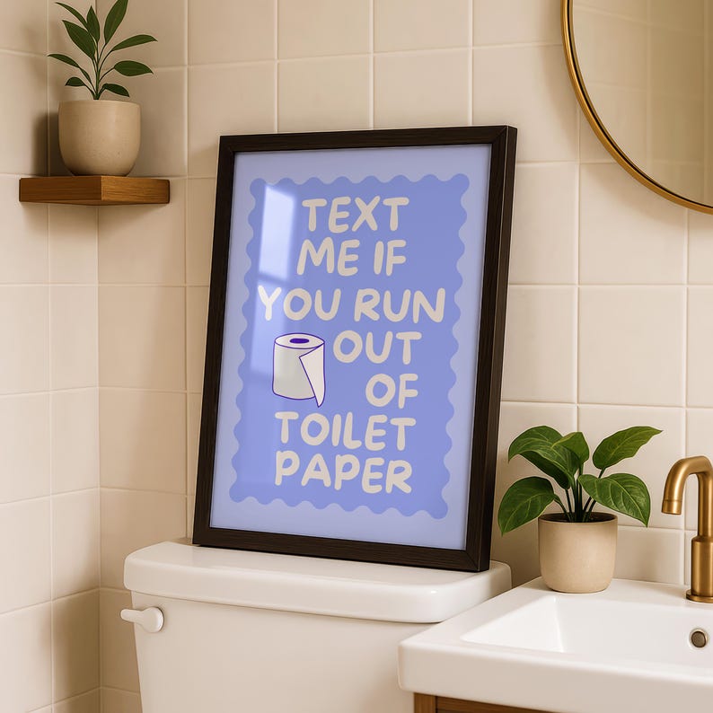 May include: A framed wall art print featuring a light blue background with a scalloped border and the white text "TEXT ME IF YOU RUN OUT OF TOILET PAPER" alongside a small illustration of a roll of toilet paper in the center of the frame.