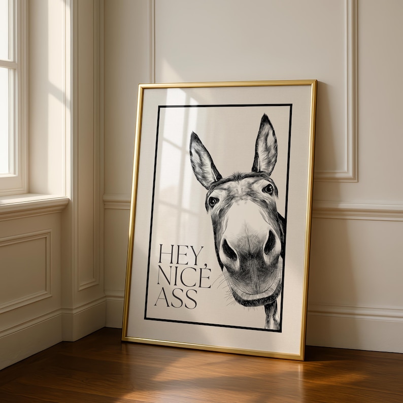 May include: A framed black and white print featuring a close-up of a donkey's face. The donkey is looking directly at the viewer. The text "HEY, NICE ASS" is printed in black below the donkey's face. The frame is gold.