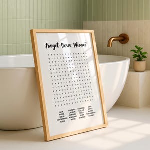 A light wood-framed print with "Forget Your Phone?" and a word search puzzle, leaning against a white bathtub, with a small potted plant on the right.