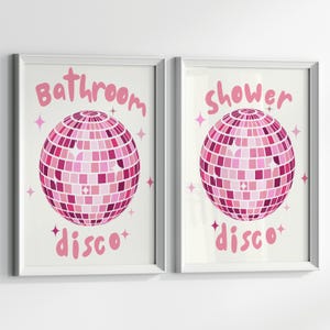 May include: Two white framed prints with pink text and a pink disco ball graphic. The text on the left print reads "Bathroom disco" and the text on the right print reads "Shower disco".