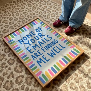 Pode incluir: Uma impress&atilde;o retangular com moldura de madeira clara. O fundo branco apresenta uma borda de barras verticais arredondadas e coloridas em rosa, amarelo, azul e verde. O texto em azul diz: NONE OF YOUR EMAILS ARE FINDING ME WELL.