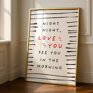 Könnte beinhalten: Ein rechteckiger gerahmter Druck mit goldenem Rand, der horizontale schwarze Linien an den Seiten und den Text NIGHT NIGHT, LOVE YOU SEE YOU IN THE MORNING in schwarzer und roter Schrift auf cremefarbenem Hintergrund in vertikaler Ausrichtung zeigt.