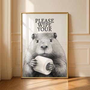 May include: A framed print with a detailed grayscale illustration of a beaver holding a roll of toilet paper. The text "PLEASE WIPE YOUR" is printed above the beaver. The artwork is set in a gold frame.