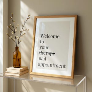 以下が含まれることがあります： ライトベージュの背景に黒い文字でWelcome to your therapy nail appointmentと書かれた額入りアートプリント。therapyという単語には取り消し線が引かれています。プリントは明るい木製のフレームに入っており、白い花を生けた茶色のガラス製花瓶の隣に置かれています。