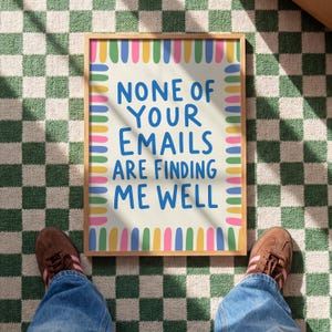 Pode incluir: Uma pe&ccedil;a de arte retangular emoldurada com um fundo creme claro e uma borda de tra&ccedil;os verticais coloridos em azul, amarelo, verde e rosa. O texto em azul diz: NONE OF YOUR EMAILS ARE FINDING ME WELL, centralizado numa fonte negrito sem serifa.