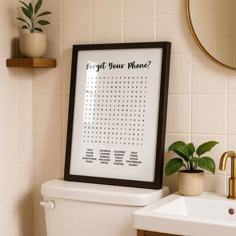 A dark wood-framed print with "Forget Your Phone?" and a word search puzzle, sitting on a white toilet tank, with a plant and a gold faucet visible.