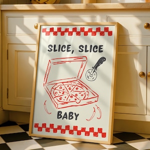 A framed print with a red and white checkered border, depicting a pizza box with a pizza inside, a pizza cutter, and the text "SLICE, SLICE BABY," leaning against white kitchen cabinets.