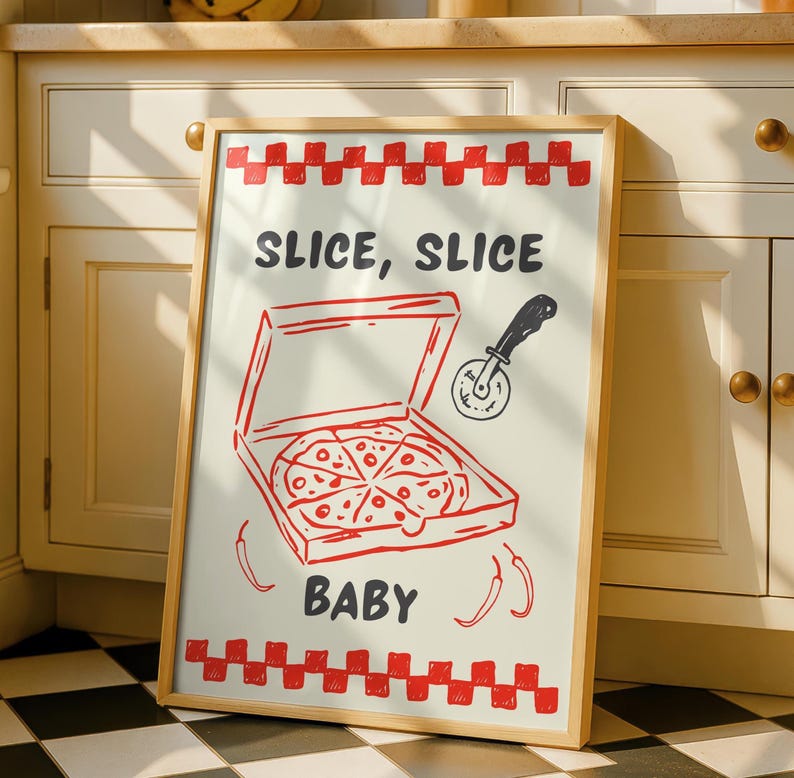 A framed print with a red and white checkered border, depicting a pizza box with a pizza inside, a pizza cutter, and the text "SLICE, SLICE BABY," leaning against white kitchen cabinets.