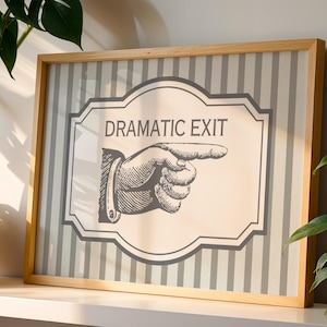 May include: A rectangular framed print featuring a light beige background with vertical gray stripes. The center displays a dark gray graphic of a pointing hand and the text DRAMATIC EXIT enclosed within a decorative border outline.