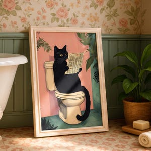May include: A framed art print depicting a black cat on a toilet, engrossed in a newspaper. The artwork is set against a pink and green backdrop, with a light beige frame. The cat has yellow eyes and a long black tail.
