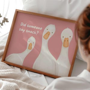 May include: A rectangular wooden frame containing a print with a dusty pink background. The print features three white geese with orange beaks looking forward. The text "Did someone say snack?" is printed in white in the upper left corner.