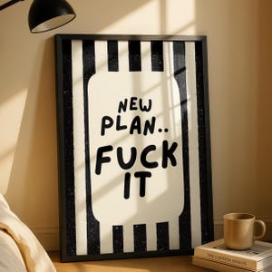 May include: A rectangular wall art print in a black frame featuring a vertical black and white striped background. A central cream-coloured rounded rectangle contains the black hand-lettered text that reads: NEW PLAN.. FUCK IT.