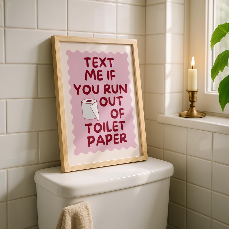 May include: A framed wall art print featuring a light pink scalloped background with dark red text that reads "TEXT ME IF YOU RUN OUT OF TOILET PAPER" and a small illustration of a white toilet paper roll centered within the text layout.