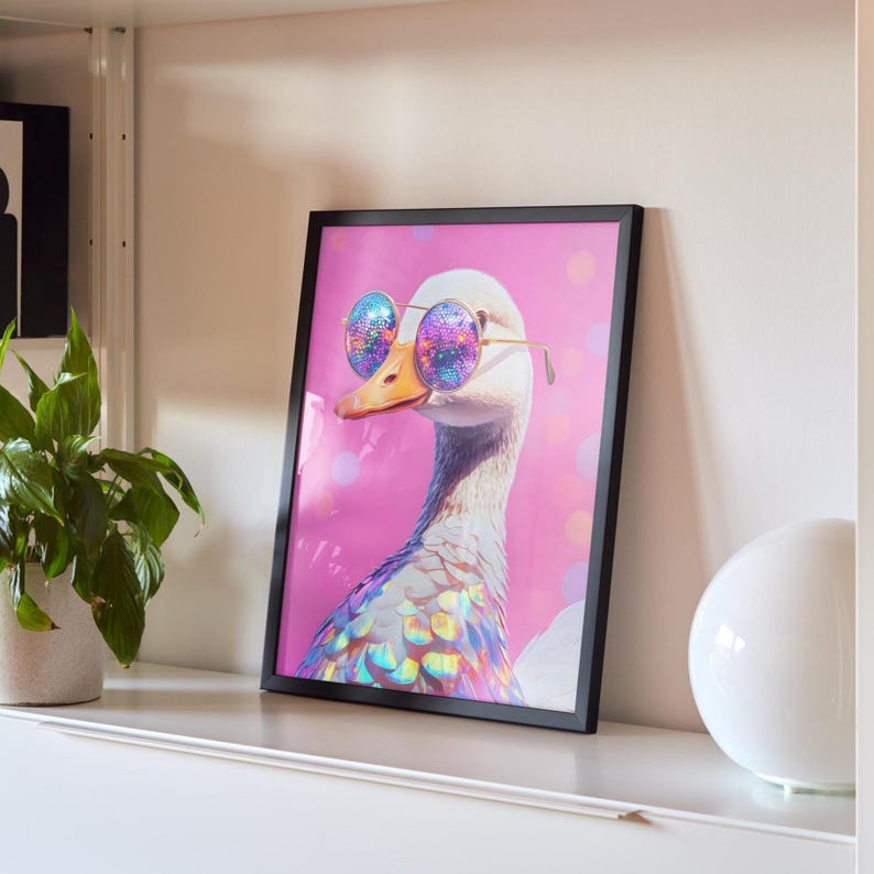 May include: A framed wall art print featuring a white goose wearing round, iridescent, multi-colored sunglasses. The goose has a metallic, rainbow-hued neck and chest, set against a vibrant pink background with soft, circular bokeh light effects.
