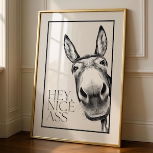 May include: Black and white donkey art print in a gold frame. The donkey is centered, with a close-up of its face. The text "HEY, NICE ASS" is printed in black, below the donkey's face.