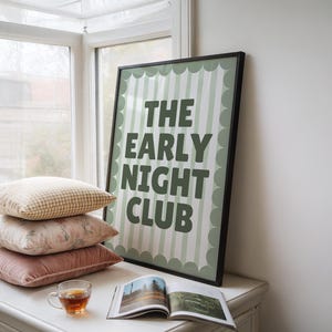 May include: A framed poster with the text "THE EARLY NIGHT CLUB" in dark green. The poster is propped against a window, next to a stack of pillows and a teacup. The poster has a light green and white striped background.