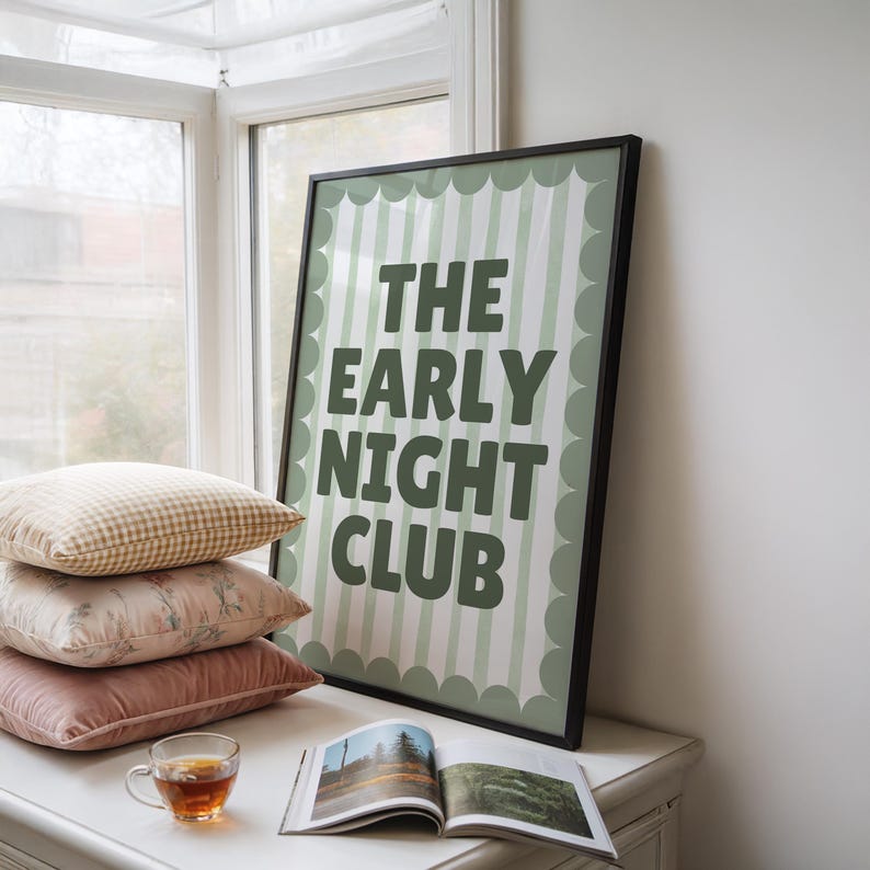May include: A framed poster with the text "THE EARLY NIGHT CLUB" in dark green. The poster is propped against a window, next to a stack of pillows and a teacup. The poster has a light green and white striped background.