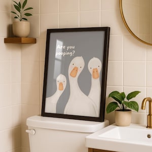 A framed print with a gray background and three white geese, with the text "Are you pooping?" above them, sits on a toilet tank in a bathroom.