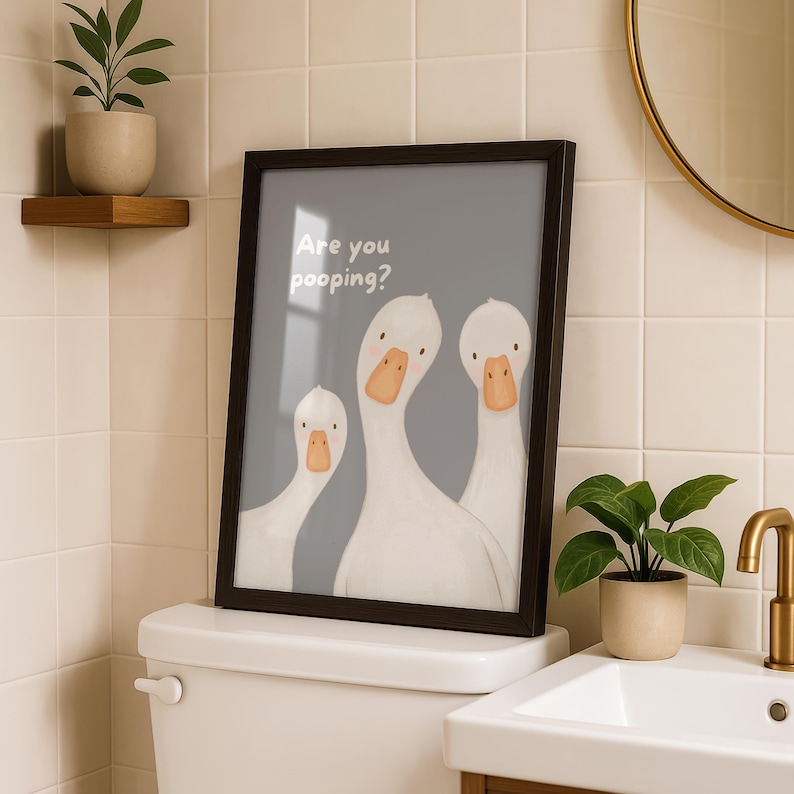 A framed print with a gray background and three white geese, with the text "Are you pooping?" above them, sits on a toilet tank in a bathroom.