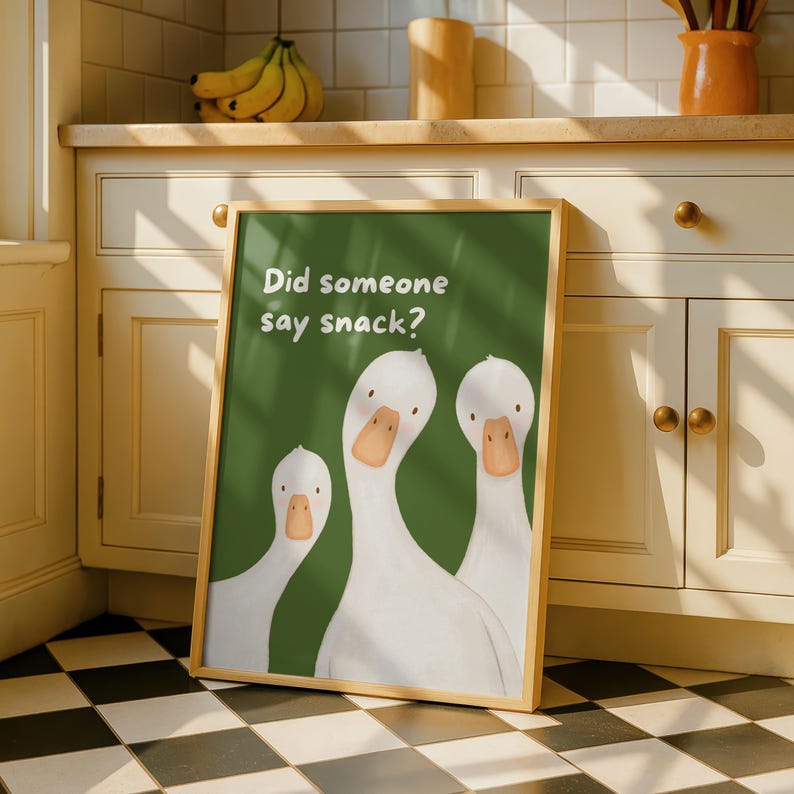 A framed print with a light wood border features three white geese on a green background, with the text "Did someone say snack?" in white, leaning against kitchen cabinets.