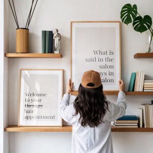 May include: Two framed wall art prints with black text on a light background. One print reads, "Welcome to your therapy hair appointment," and the other reads, "What is said in the salon stays in the salon." The prints are displayed on wooden shelves.
