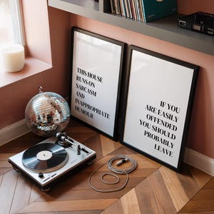 May include: Two rectangular black-framed wall art prints with white backgrounds and black text. The left print reads: THIS HOUSE RUNS ON SARCASM AND INAPPROPRIATE HUMOUR. The right print reads: IF YOU ARE EASILY OFFENDED YOU SHOULD PROBABLY LEAVE.