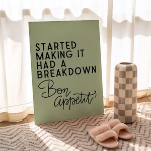 Peut inclure: Une affiche vert sauge avec le texte "STARTED MAKING IT HAD A BREAKDOWN Bon appétit!" est posée sur un tapis à motifs. Un vase à carreaux et des pantoufles roses sont à côté de l'affiche. La pièce a une palette de couleurs neutres.