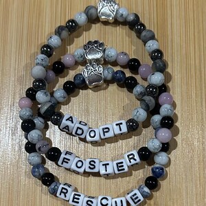 May include: Three beaded bracelets with the words "ADOPT", "FOSTER", and "RESCUE" spelled out in white letter beads. The bracelets feature a mix of black, gray, and pink beads, and are accented with silver paw print charms.