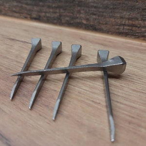 May include: Five silver horseshoe nails are arranged on a wooden surface. The nails are made of metal and have a rectangular head and a pointed tip. These nails are used to attach horseshoes to a horse's hoof.